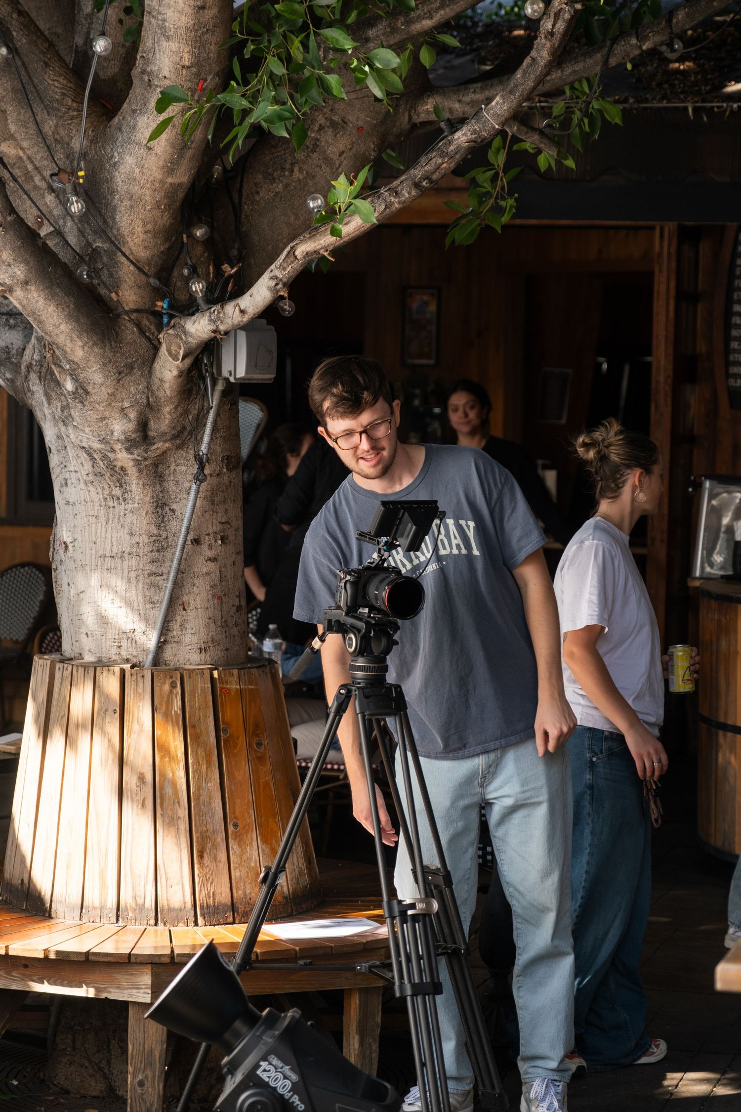 Film crew setting up camera equipment under a large tree during a video production shoot.