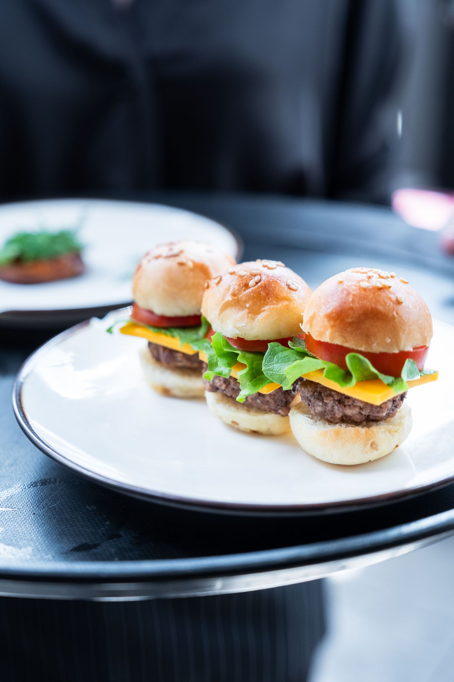 Four miniature cheeseburger sliders with lettuce on white plates against a dark background.