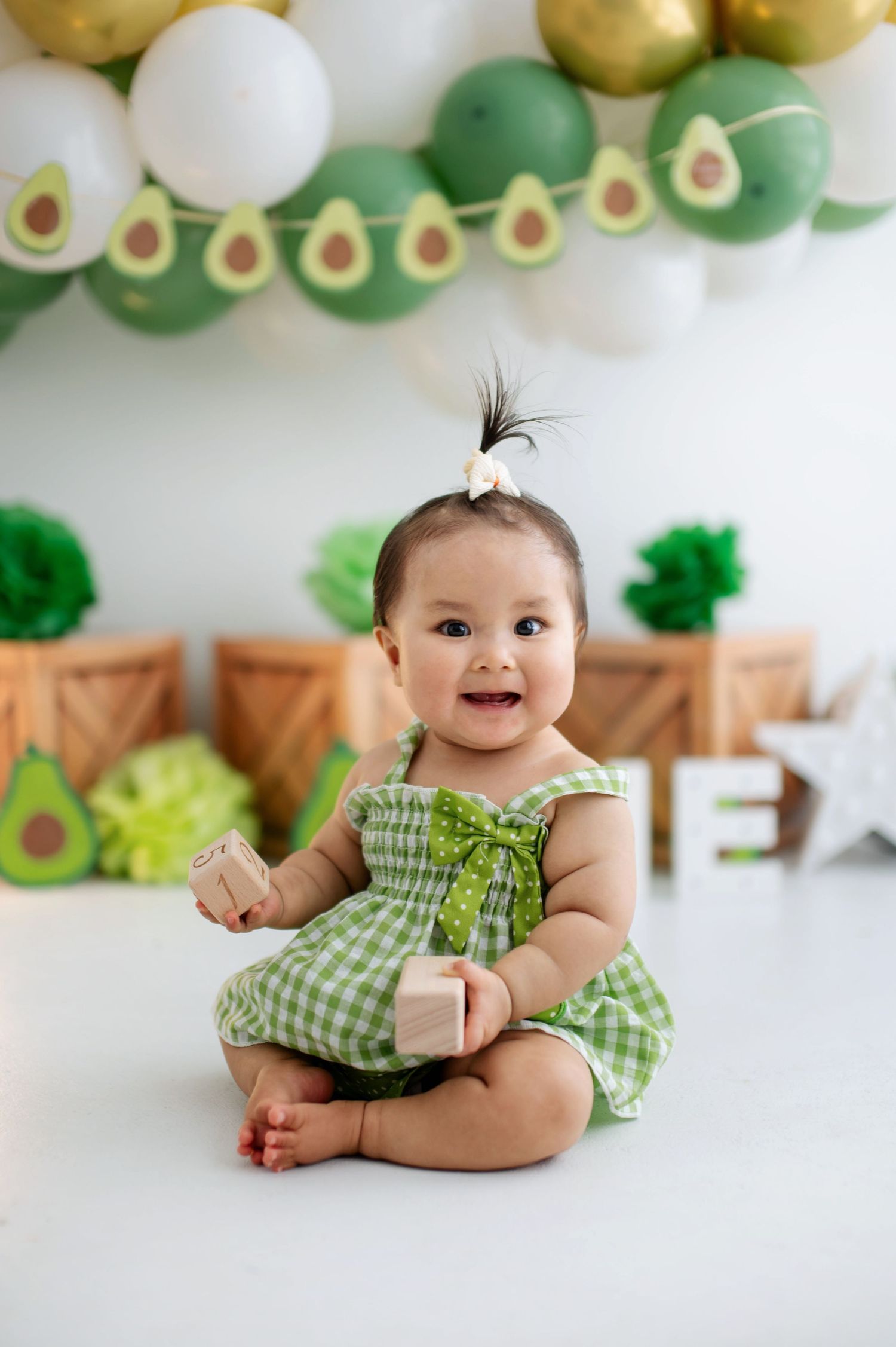 Baby wearing green gingham dress sits on white floor with avocado-themed balloon garland and party decorations.