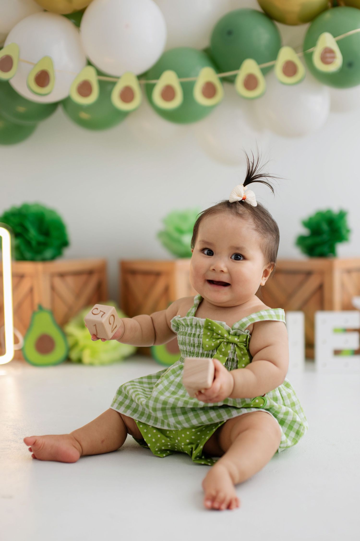 Baby in green checkered dress smiles while sitting against wall decorated with avocado and balloon garland.