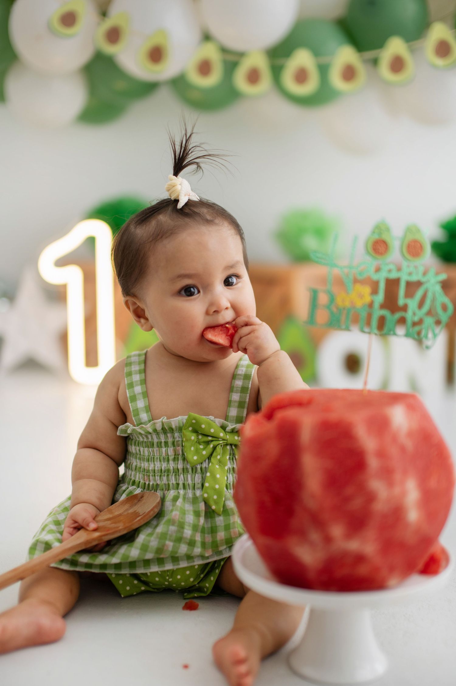 Baby in green dress reaches for red birthday cake with number 1 light and avocado-themed decorations.