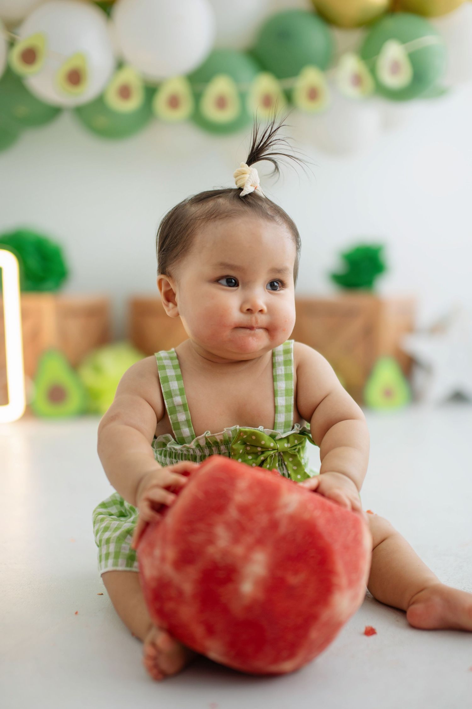 Baby in green checkered outfit sitting with large watermelon against festive avocado-themed party background.