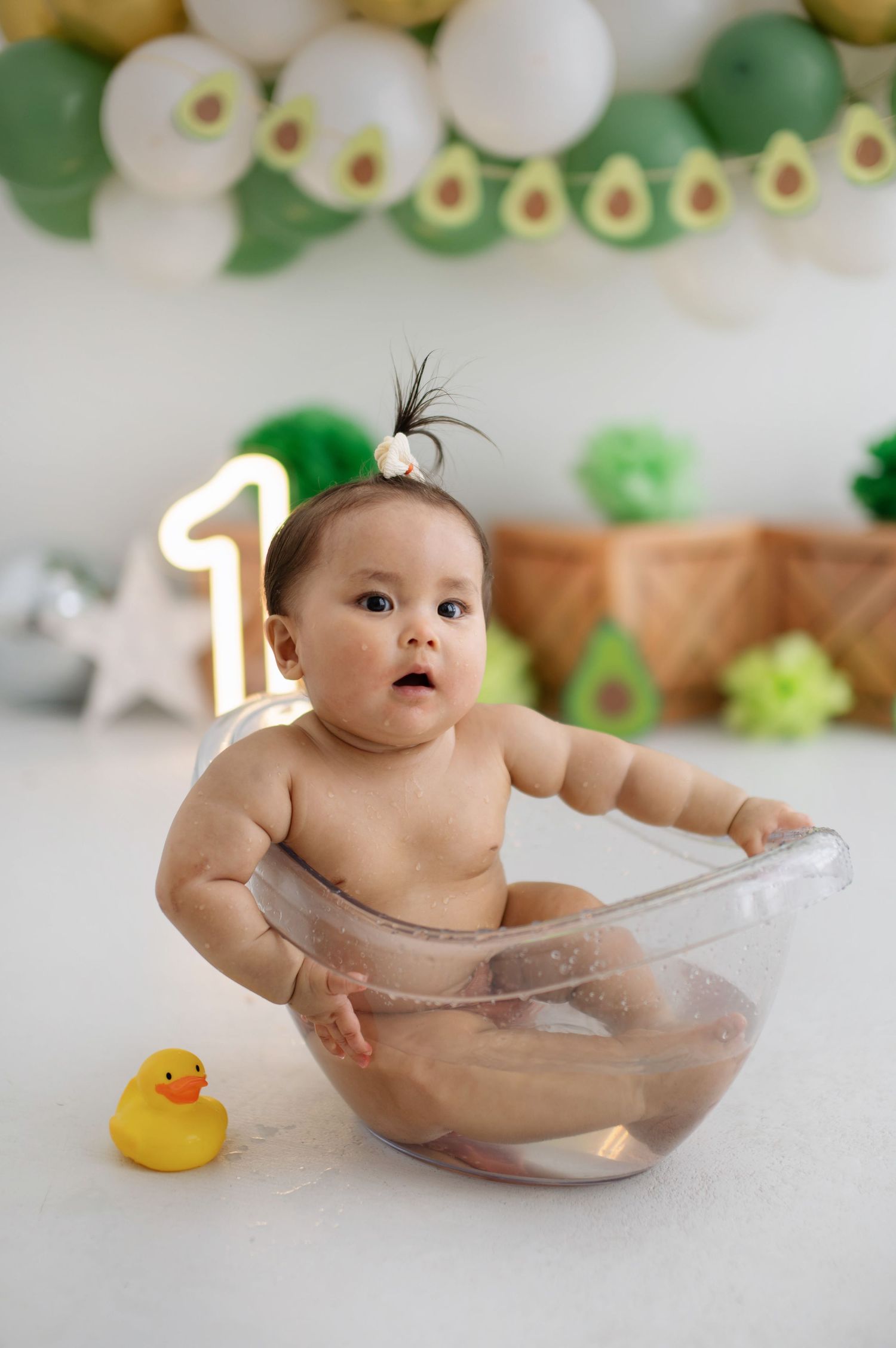 Baby sitting in clear bathtub with rubber duck during avocado-themed first birthday photoshoot.
