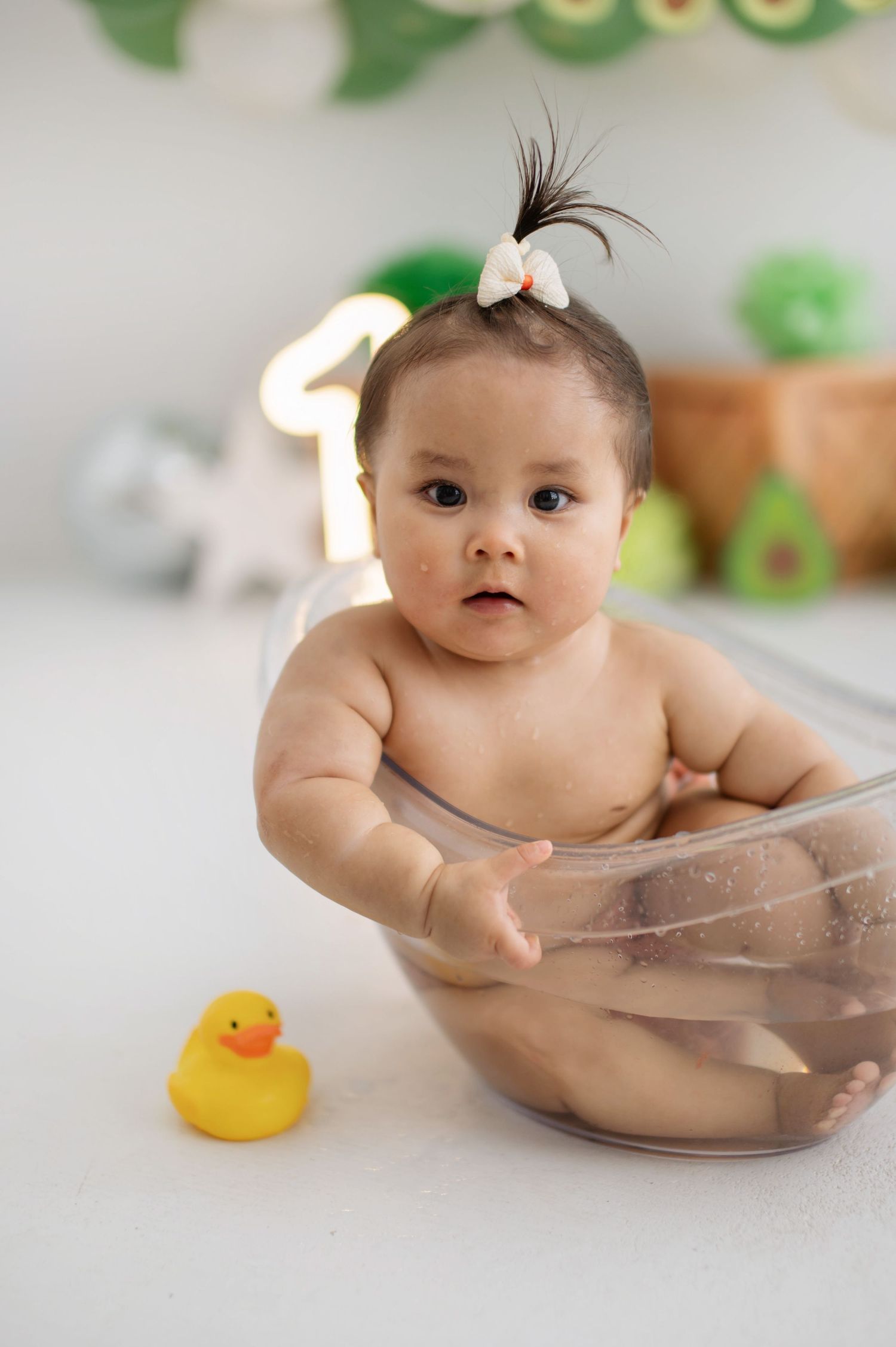 A baby sits in a clear plastic tub with a yellow rubber duck toy during bath time.