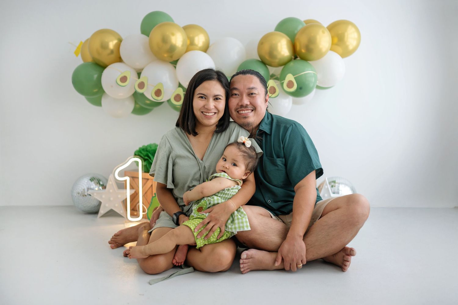 Family poses together on white floor with green and gold balloon arch and decorations for special occasion photos.