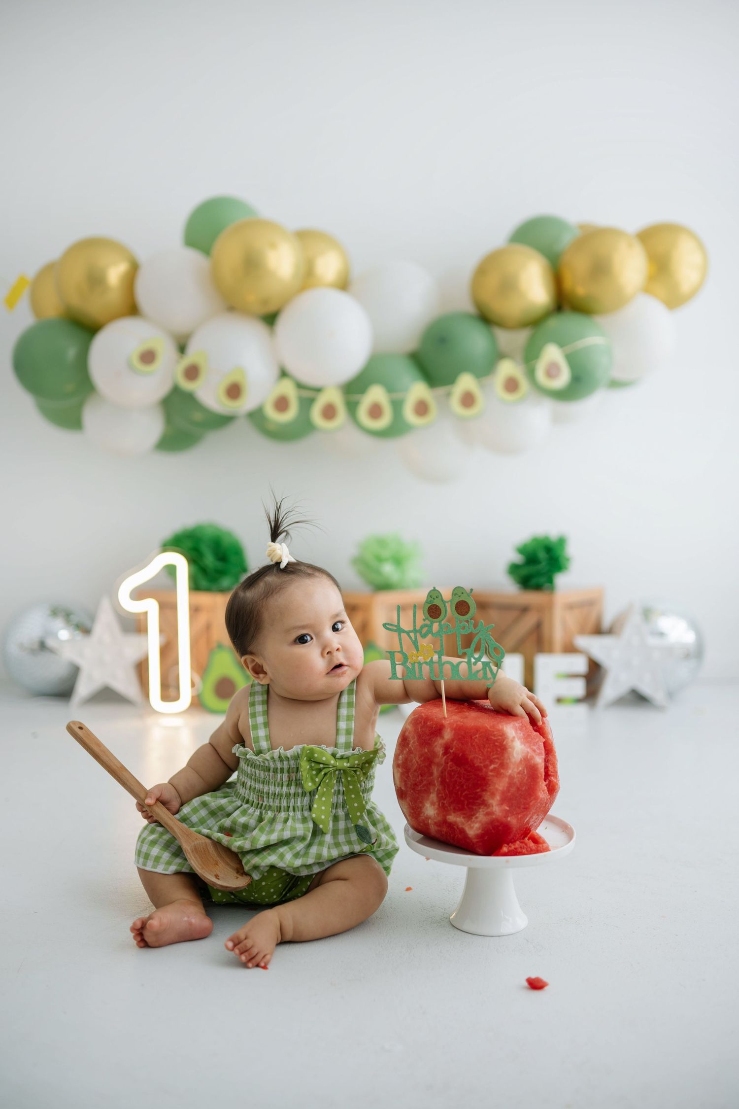 Baby in green gingham outfit lying next to red watermelon cake during first birthday photoshoot.