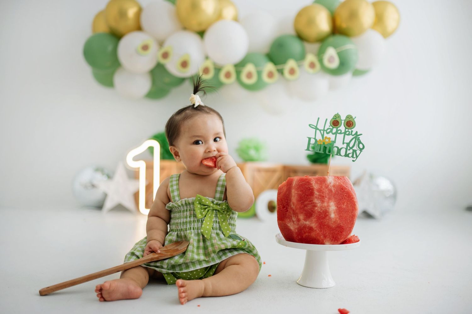 A baby in a green dress sits on the floor with watermelon and party decorations in a bright white studio setting.