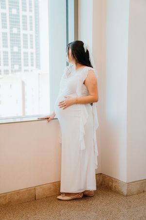A person in a flowing white dress stands near a window in a minimalist interior space with white walls.