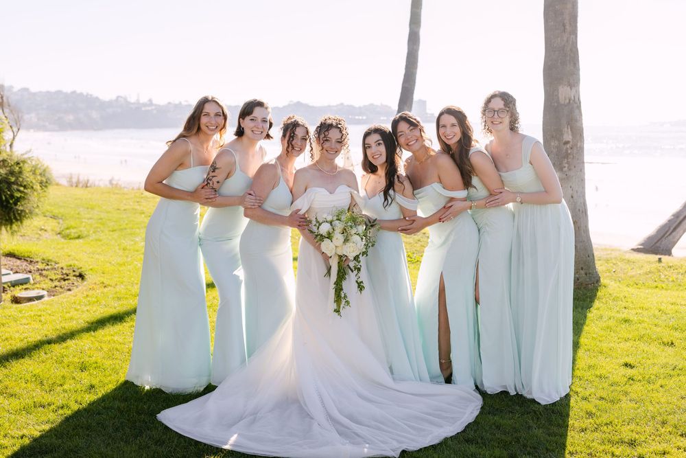 A wedding party in light blue bridesmaid dresses poses outdoors on a sunny day by the water.