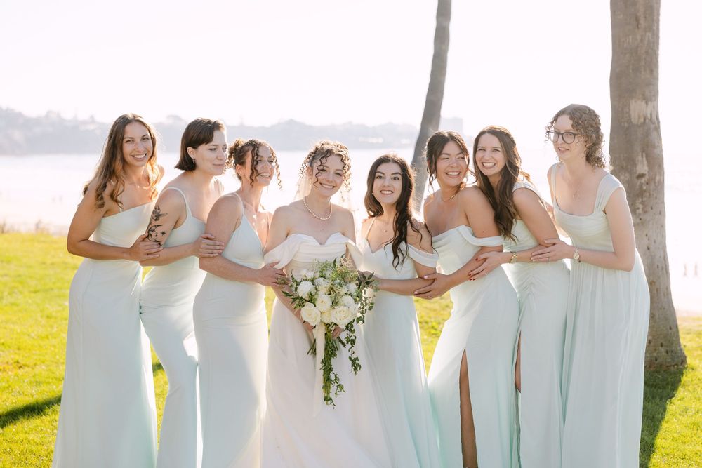 Wedding party in mint green dresses poses together in sunlit outdoor setting with bouquets.