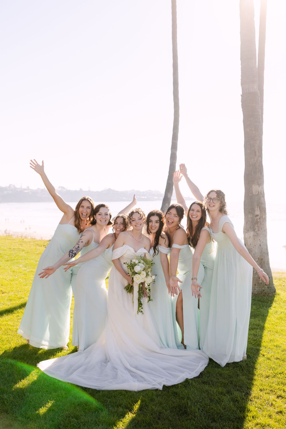 Bridal party in mint green dresses and white gown pose joyfully among palm trees at sunset.