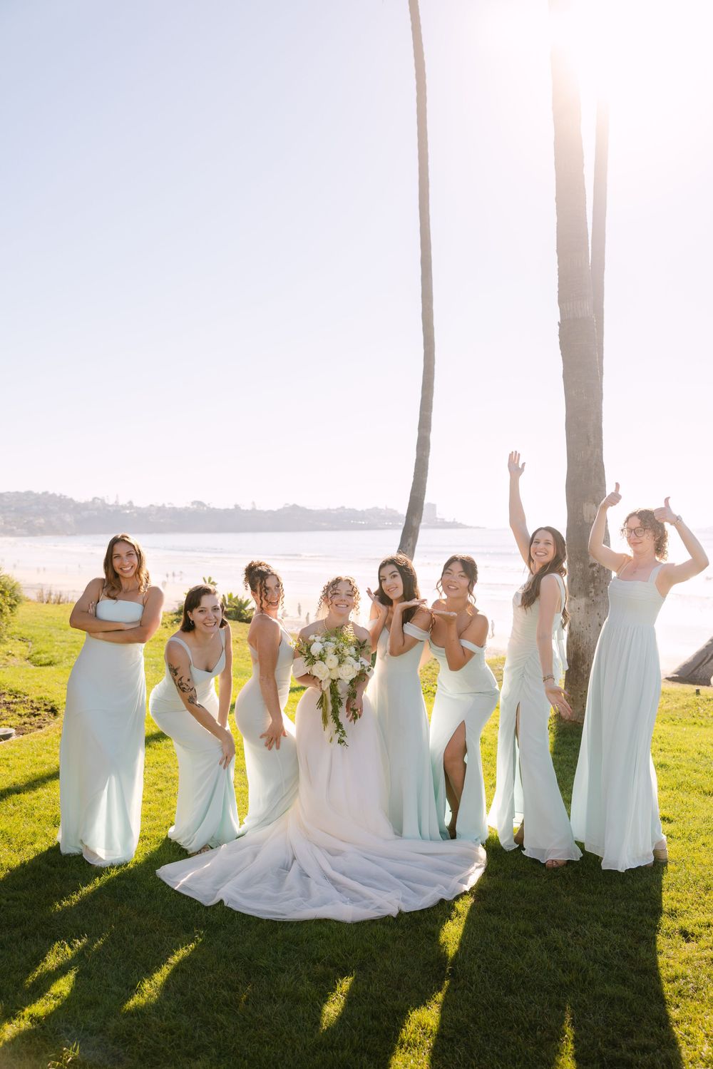 Bridal party in white dresses poses together on grassy lawn with sunlight streaming through trees behind them.