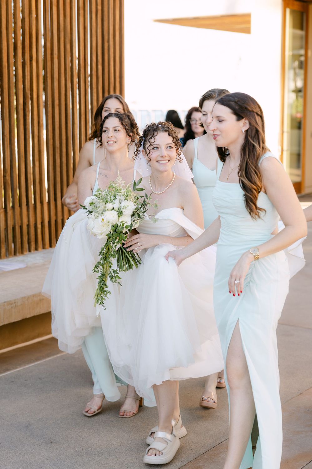Bridesmaids in light blue dresses and white attire laugh and dance together in a covered outdoor walkway.