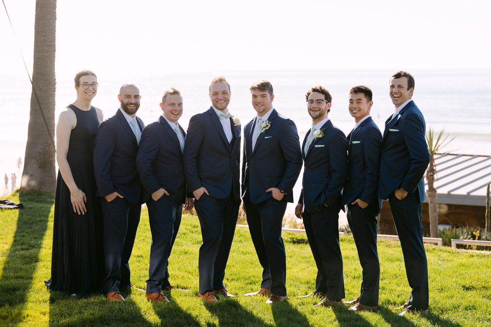 Wedding party in black suits stands in line on grassy hillside with ocean view in background.