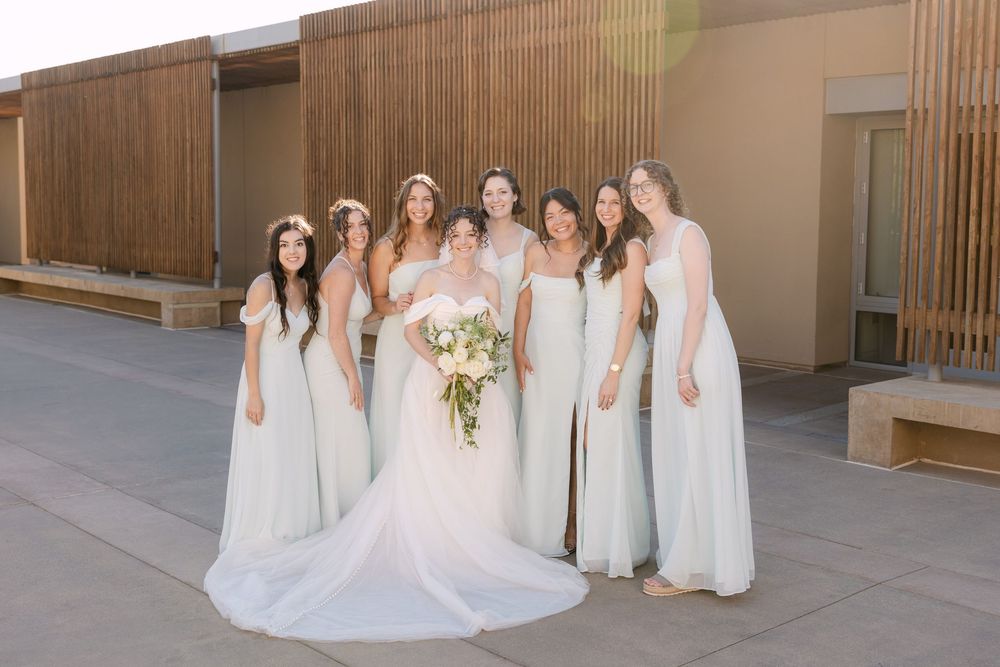 A wedding party wearing white dresses poses together in a wooden-paneled venue hallway.