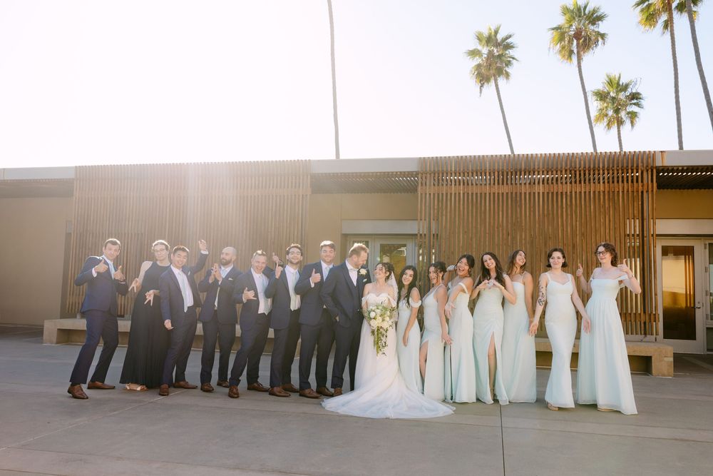 Wedding party in light blue dresses and dark suits posing in front of palm trees and modern building.