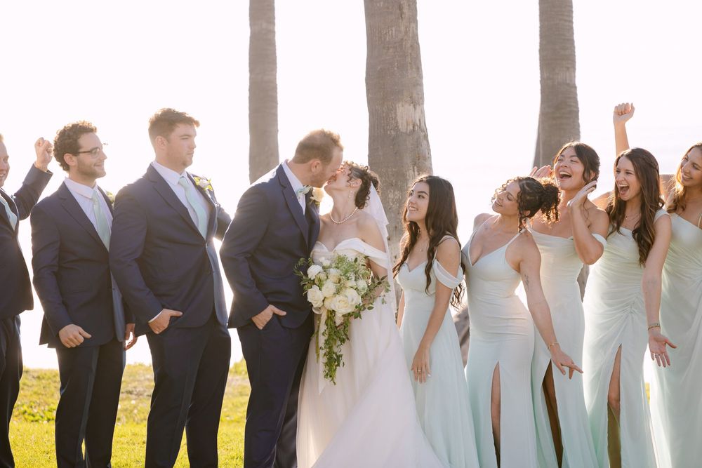 Wedding party in formal attire laughing together outdoors with sunlit trees in background.
