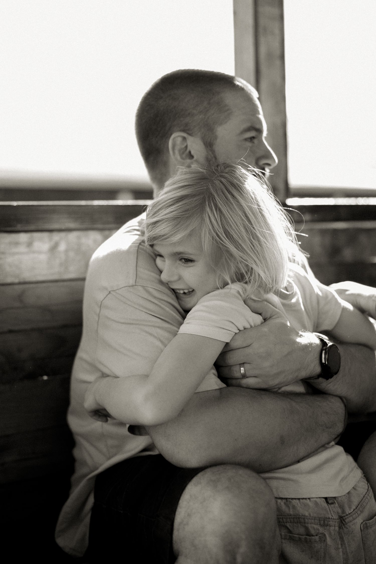 Black and white photo of a man hugging a young blonde girl who smiles warmly, seated near a window.