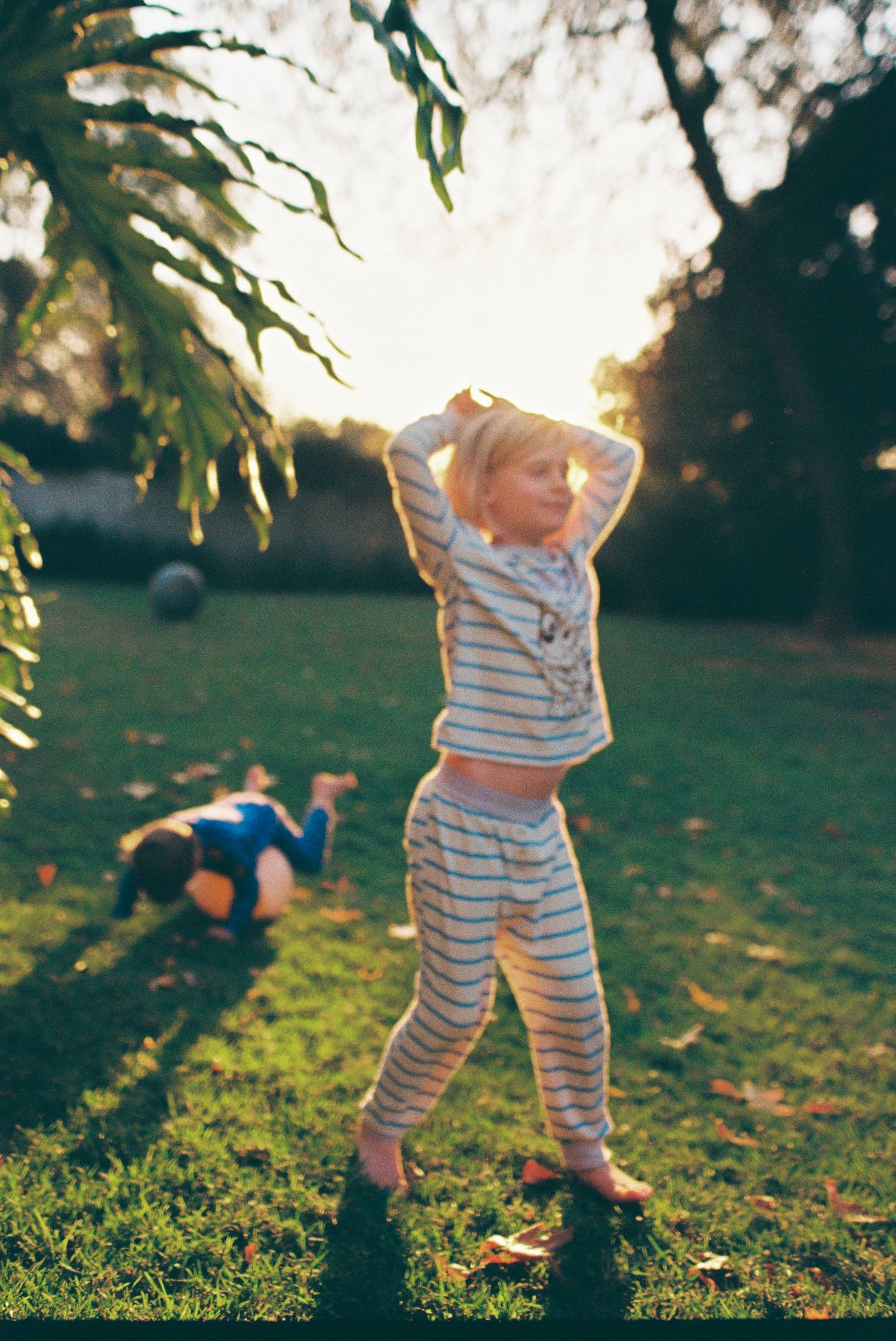 A young child in striped pajamas plays outdoors in a sunlit garden at golden hour, with another child playing in the background.