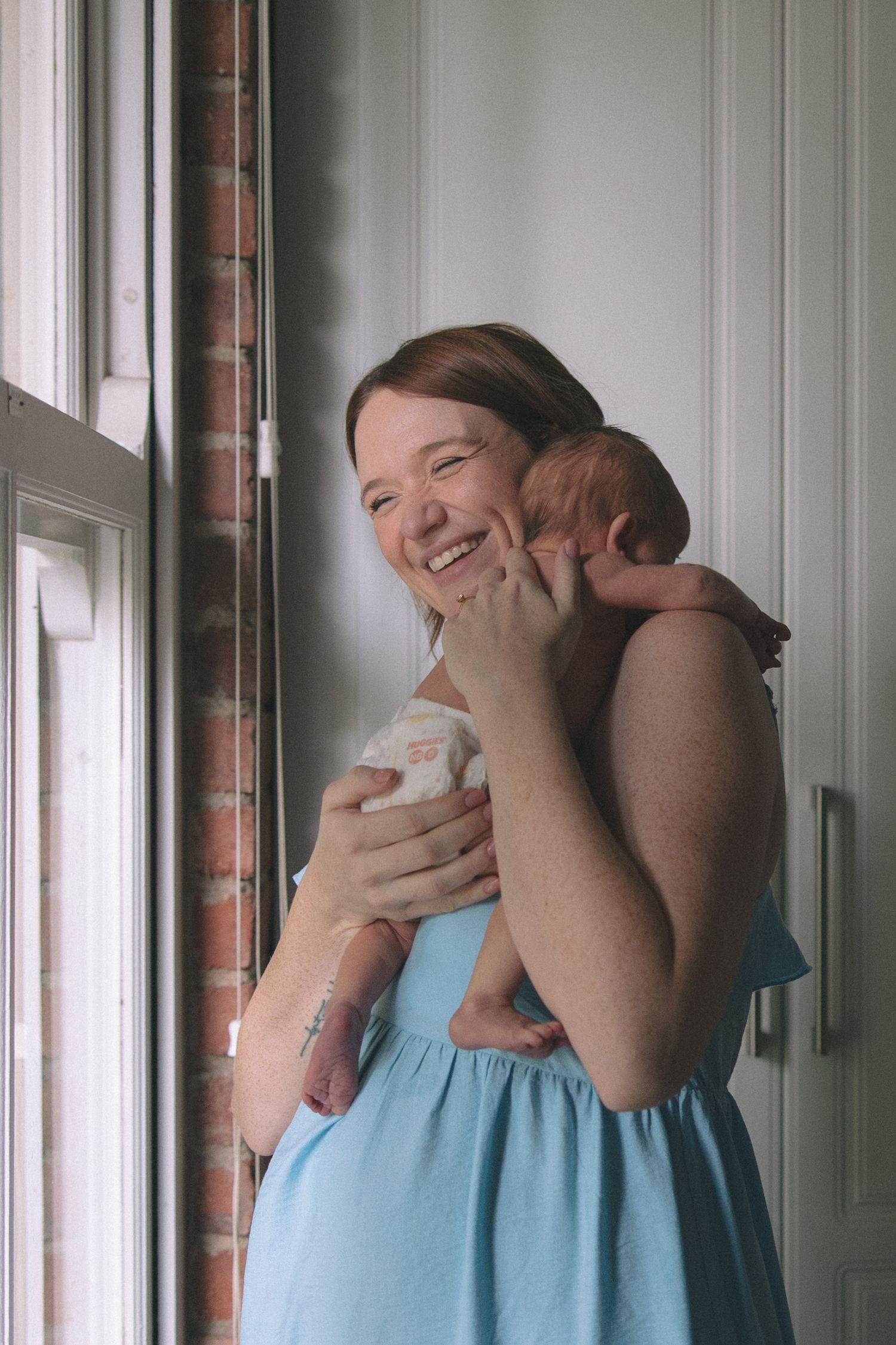 Smiling mother in blue dress holds newborn baby near window with brick wall background.