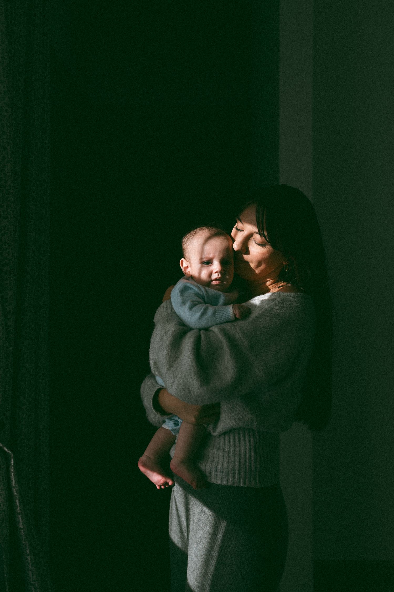 A mother tenderly holds her baby in soft natural light against a dark background, both dressed in cozy gray clothing.