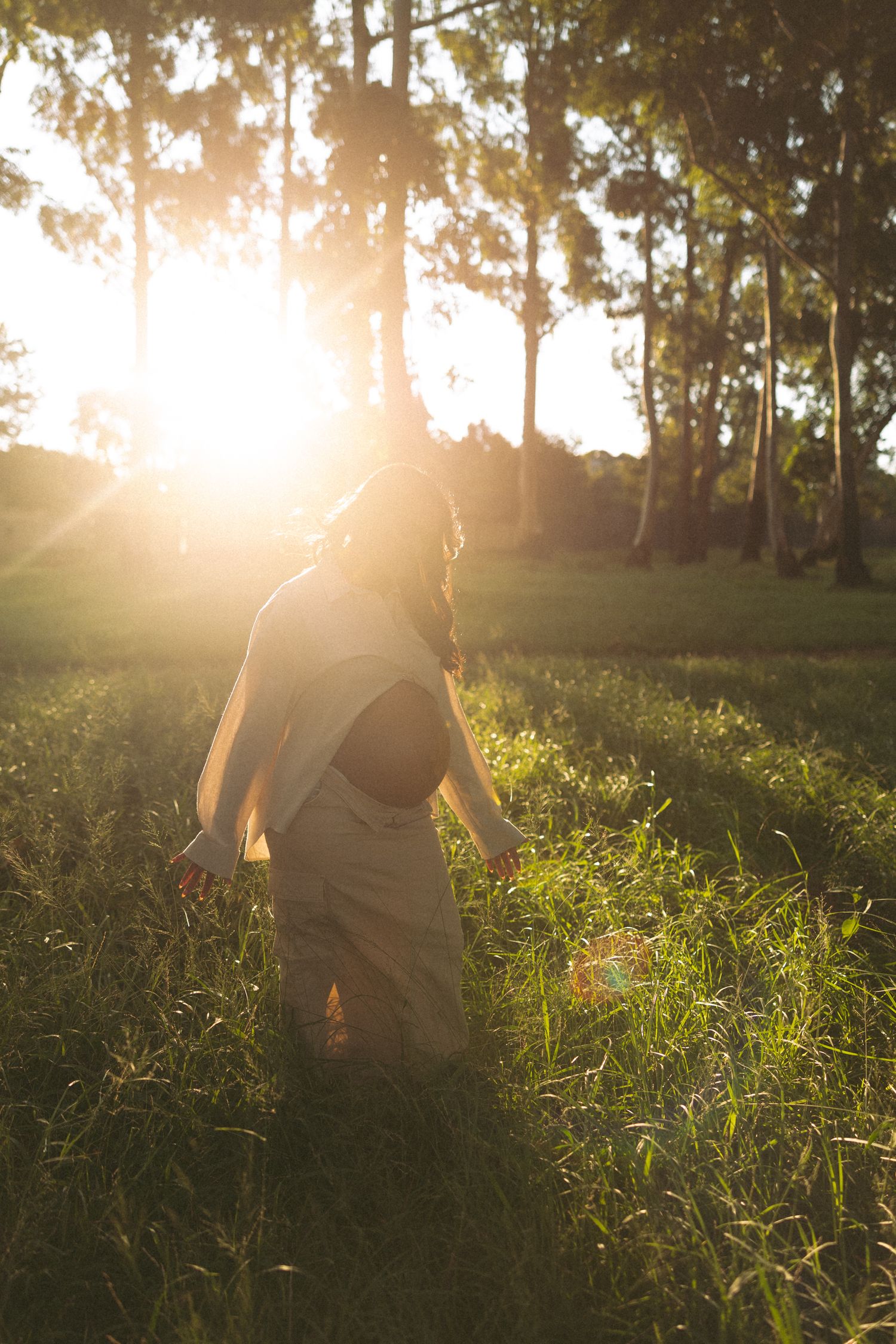 Woman in dress walking through sunlit green meadow with tall trees and golden sunset light in background.
