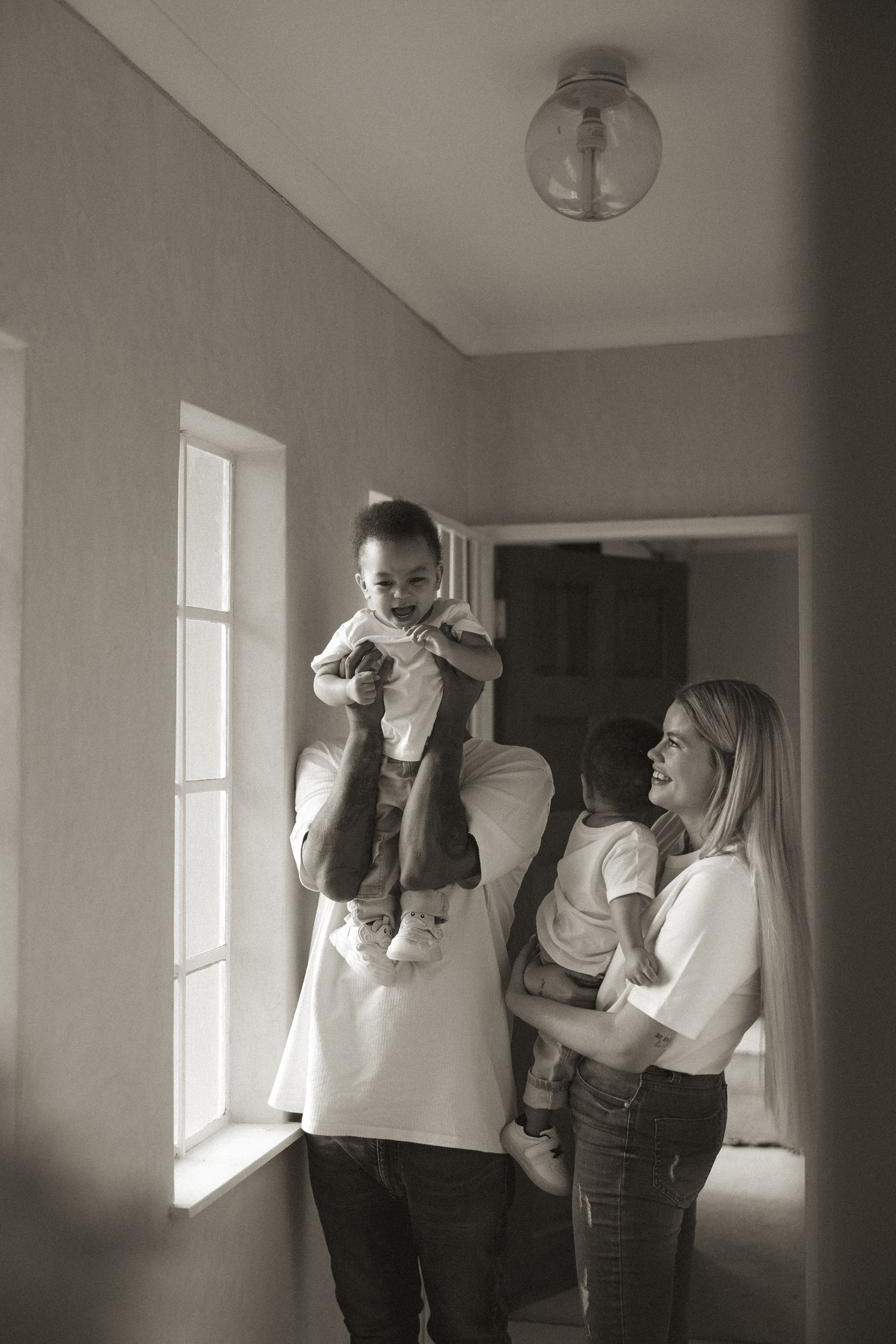 Black and white photo of a happy family with parents holding two young children near a sunlit window indoors.