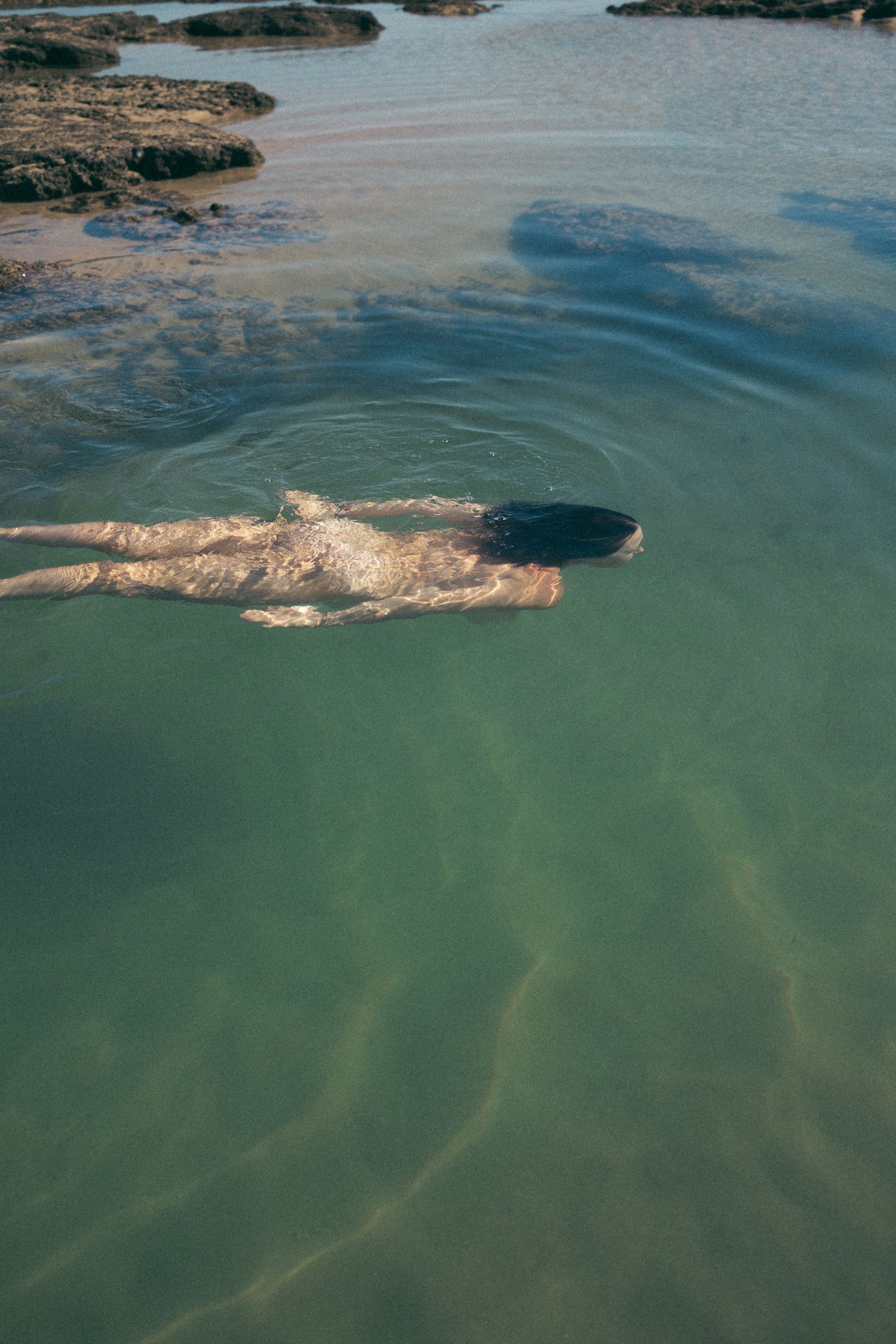 Person swimming in clear turquoise ocean water near rocky shoreline, viewed from above.