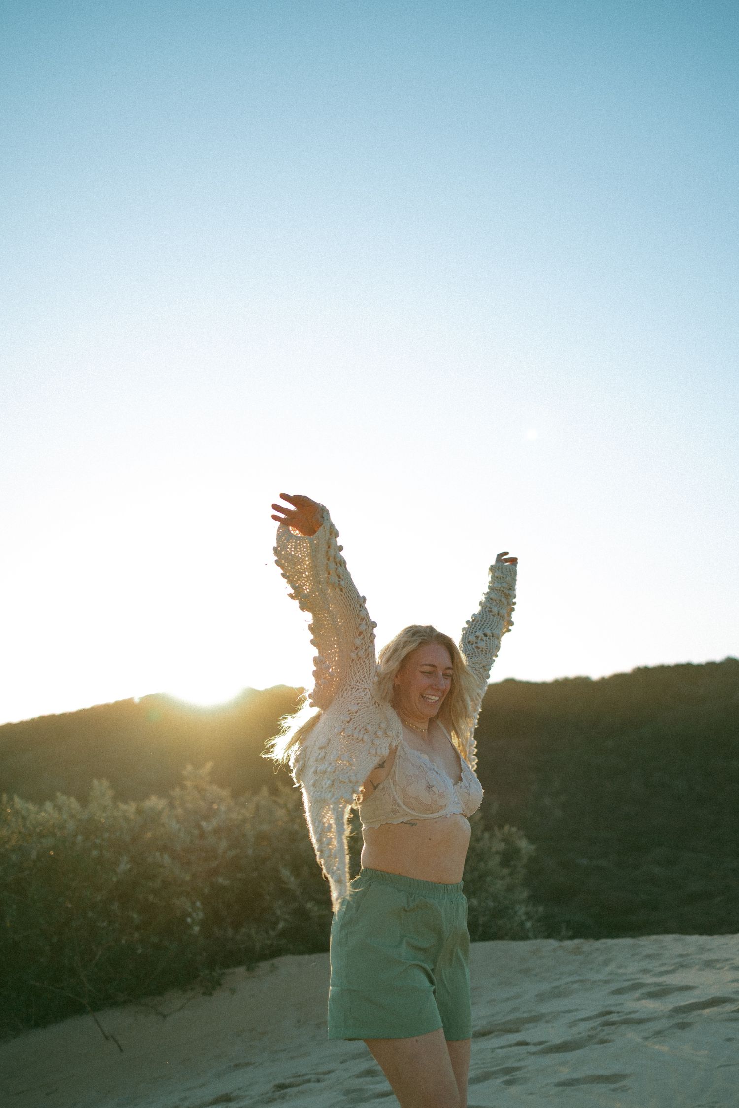 Joyful blonde woman in green shorts and fringed top dancing on sand dunes at golden sunset.
