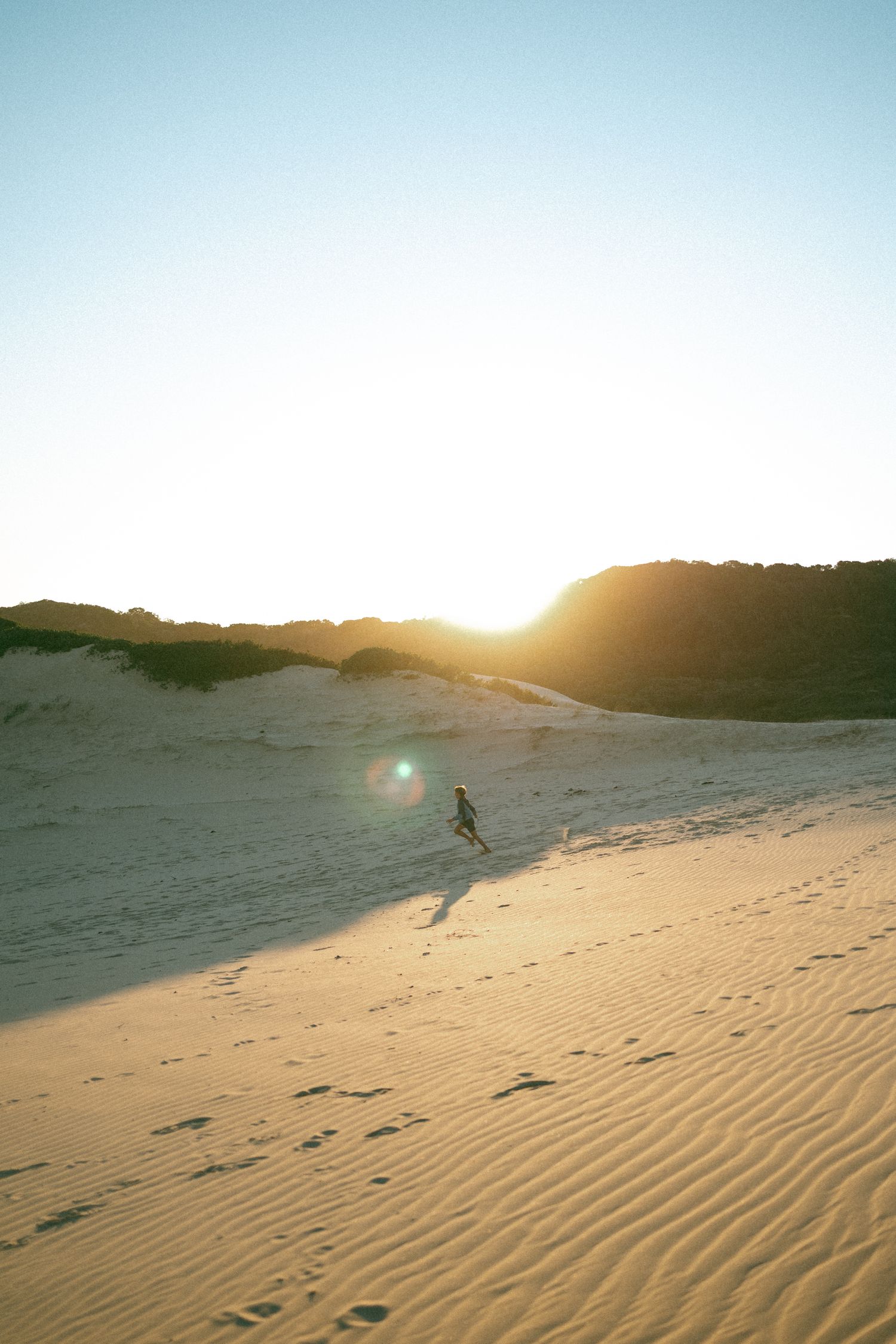 Person running on golden sand dunes at sunset with lens flare and rippled sand patterns in a vast desert landscape.