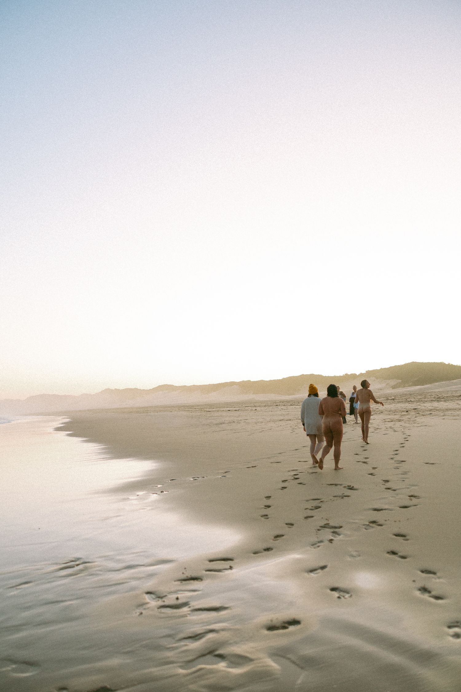 Group of friends walking along a sandy beach at sunset, leaving footprints in the sand near the shoreline.