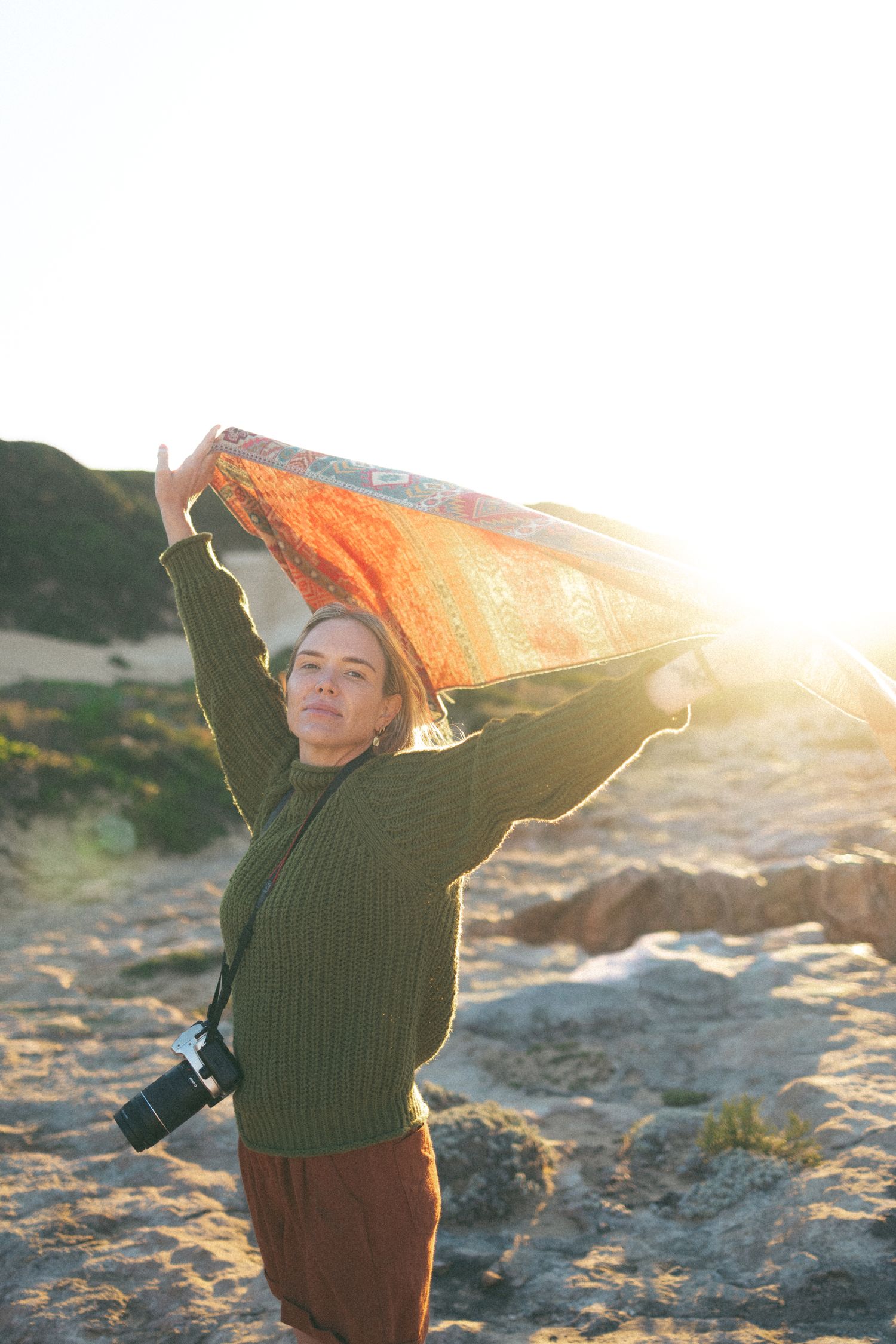 Woman in green sweater holds orange scarf overhead on sandy beach at golden sunset with camera around neck.