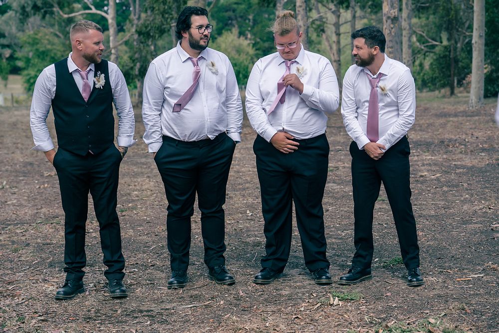 A group of groomsmen in formal attire pose casually outdoors in a wooded setting.