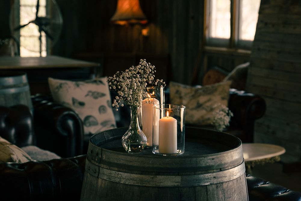 White pillar candles and baby's breath flowers arranged on rustic wooden barrel in cozy dark interior with ambient lighting.
