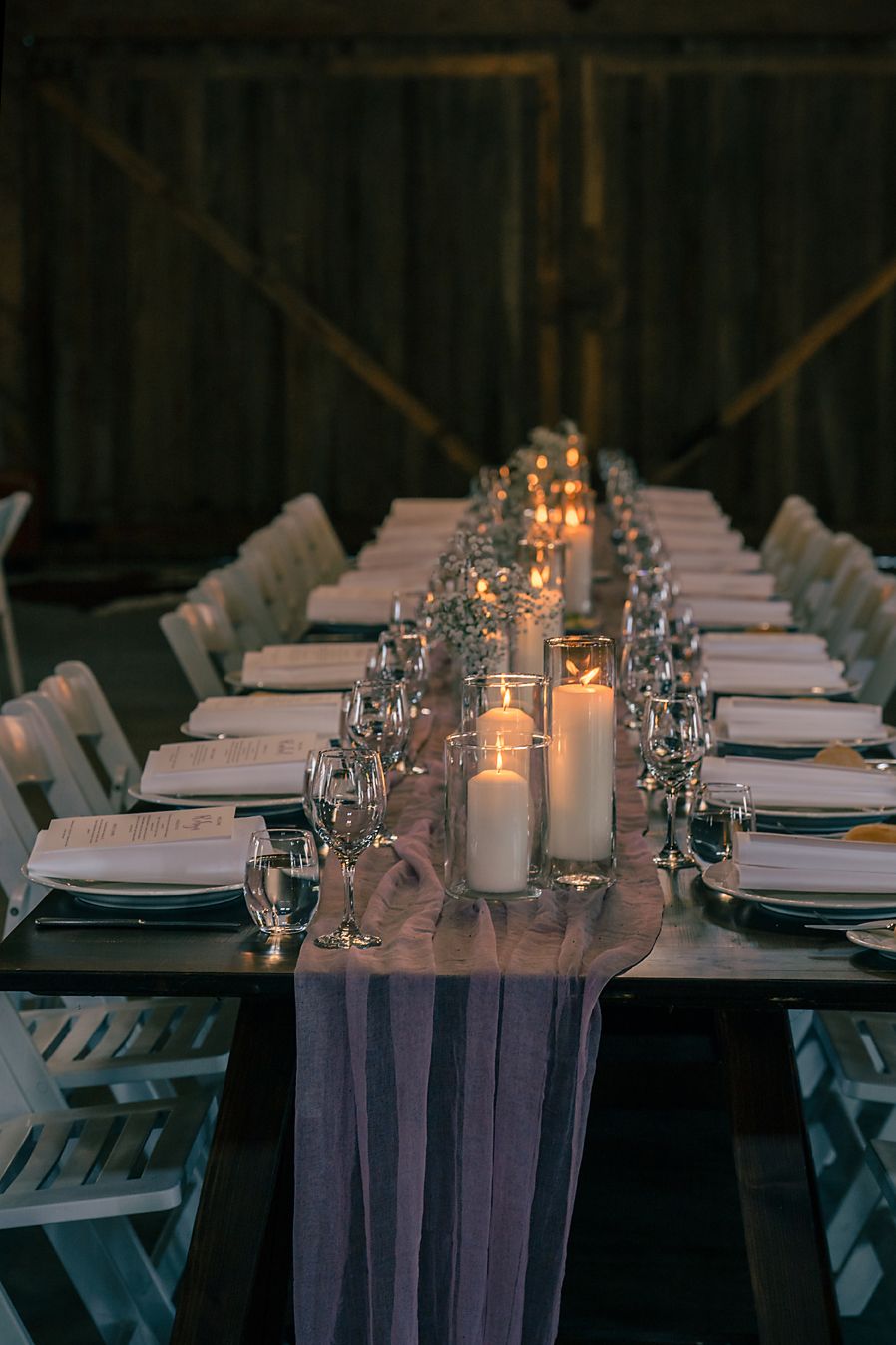 Elegant wedding reception table settings with glowing candles and white linens in a rustic barn venue.