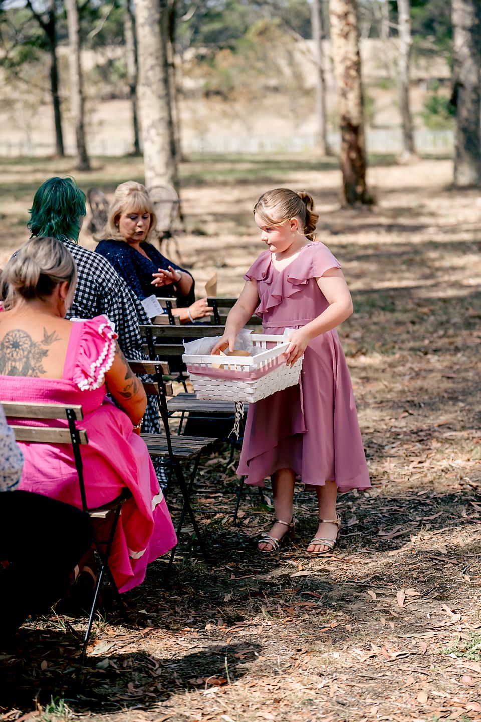 People in pink dresses and casual attire gather for a picnic in a wooded outdoor setting.