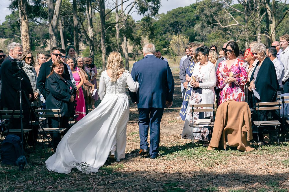 Wedding party and guests gather outdoors in a natural wooded setting during a fall celebration.