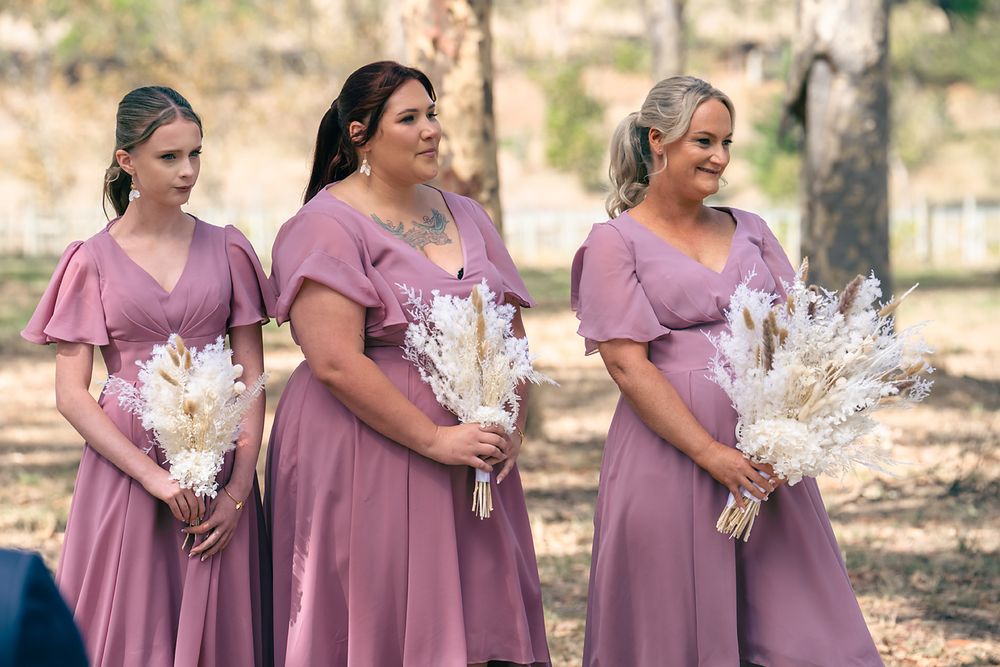 A group wearing matching mauve dresses stands together holding white floral bouquets outdoors.