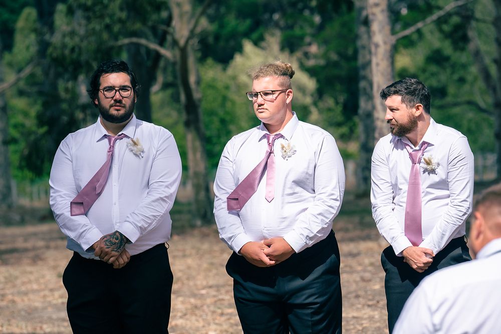 Three groomsmen in white dress shirts and pink ties stand together during an outdoor wedding ceremony.