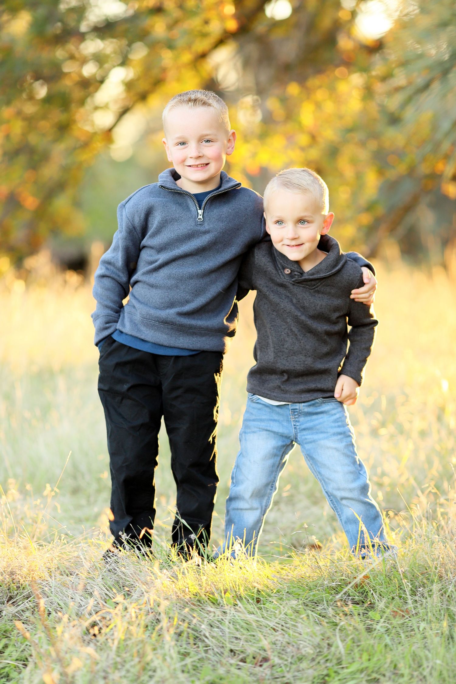 Two children in casual clothing stand together in a golden autumn field during sunset.