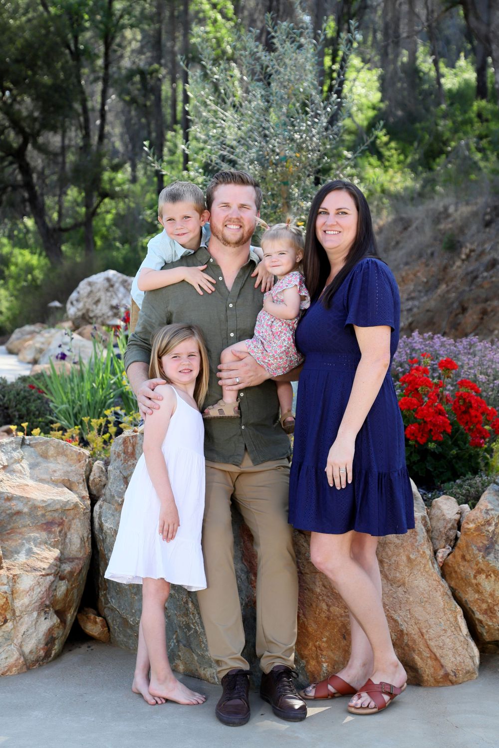 A family poses together in front of a landscaped garden with colorful flowers and large rocks on a sunny day.