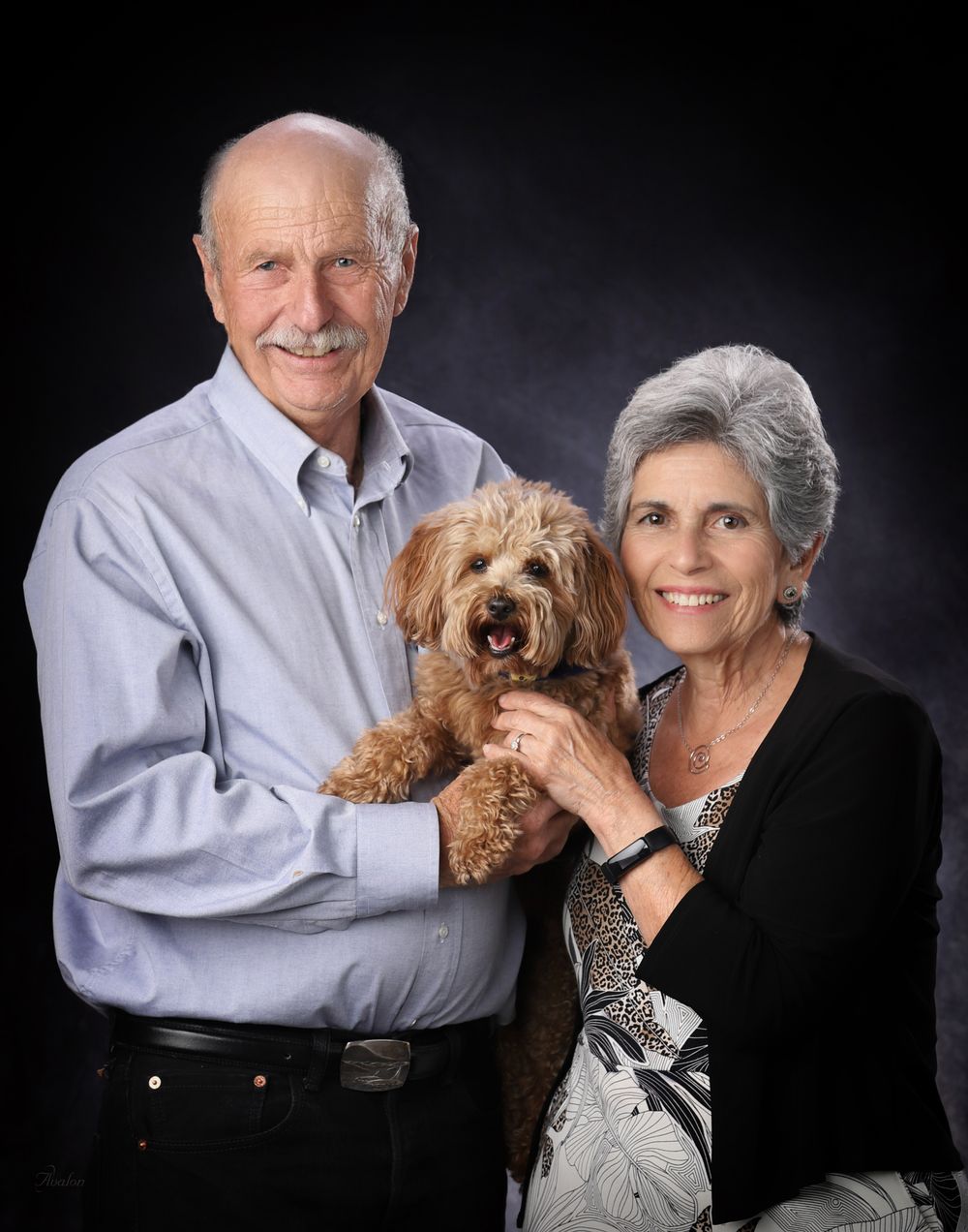 Elderly couple poses with their small brown dog in a professional studio portrait series against dark background.