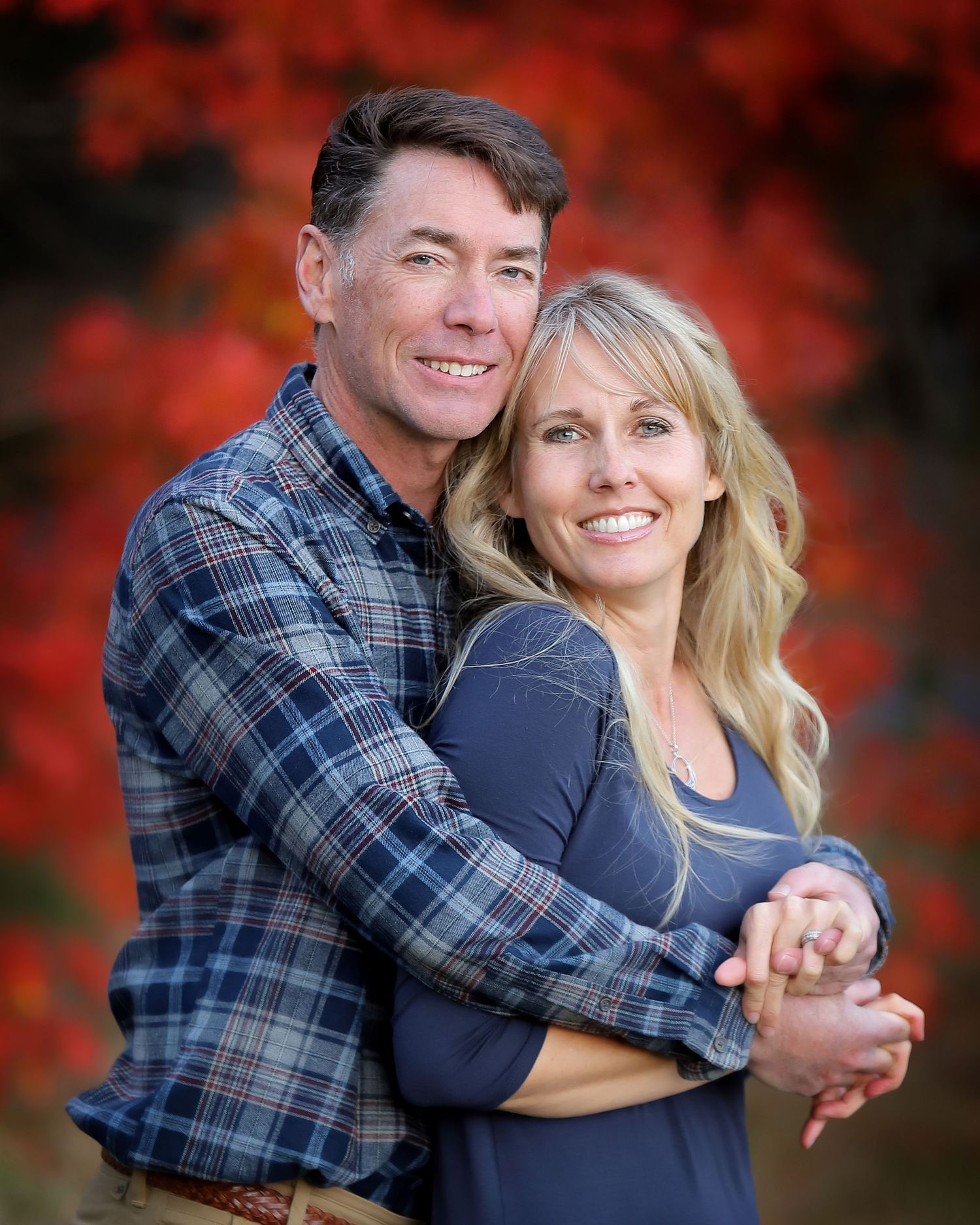 Couple embraces while posing against vibrant red autumn foliage backdrop during outdoor photo session.