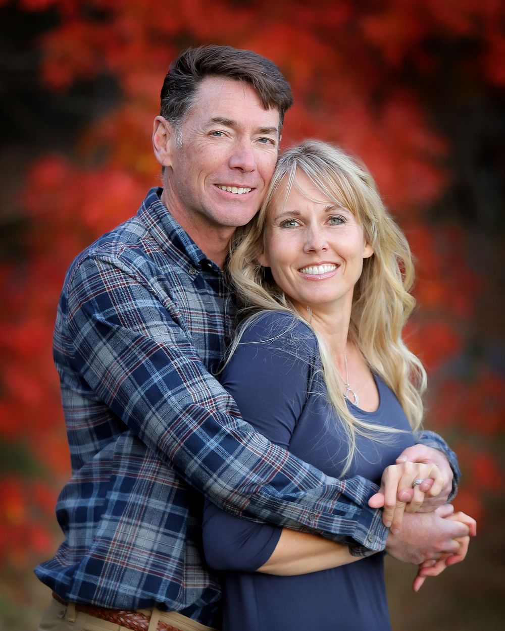 Couple embraces while posing against vibrant red autumn foliage backdrop during outdoor photo session.