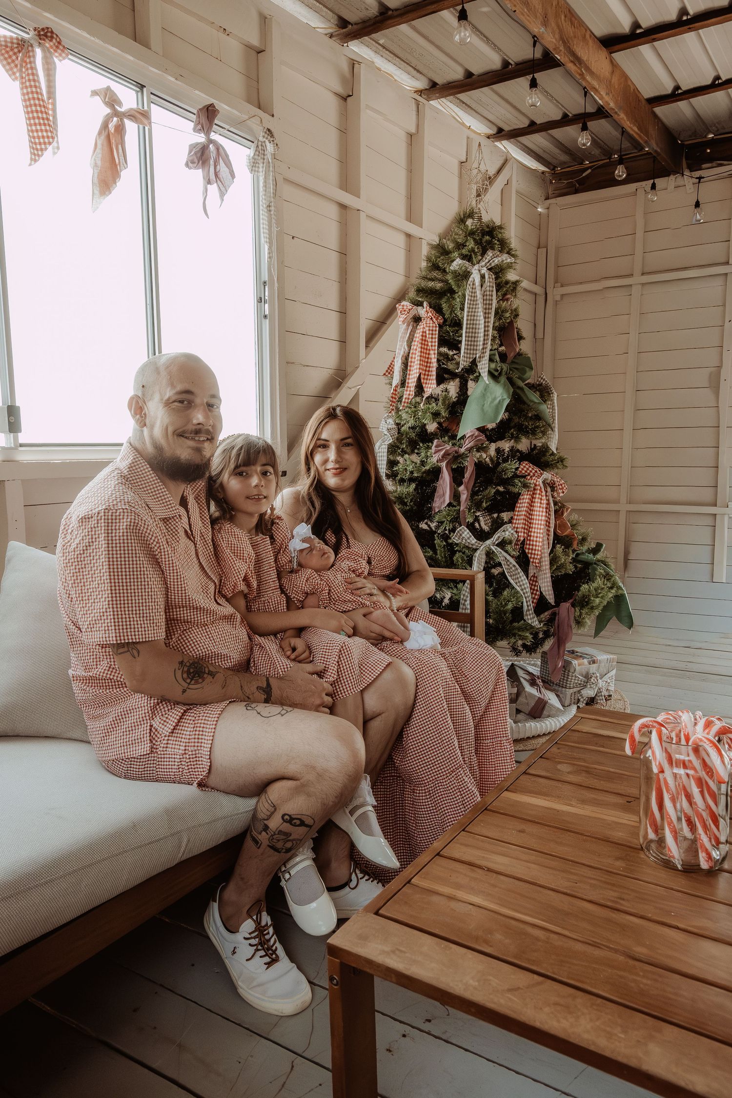 Family in matching outfits sitting on bench in rustic room with holiday decorations at a central coast christmas photoshoot.