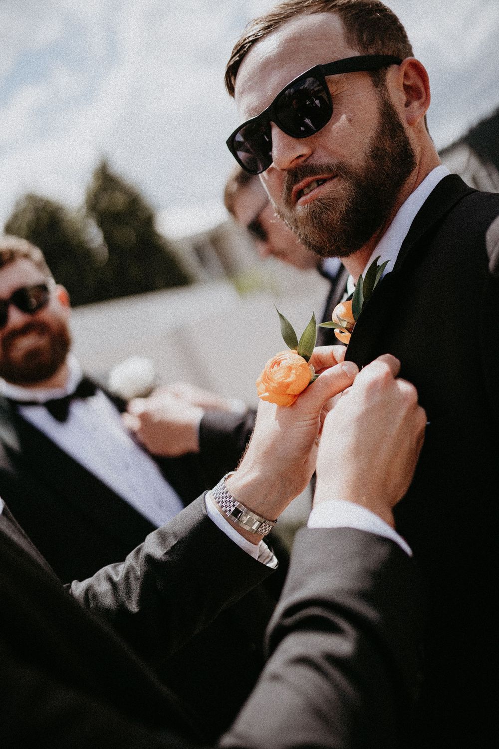 Groomsmen in sunglasses adjusting their boutonnieres against a bright sky.