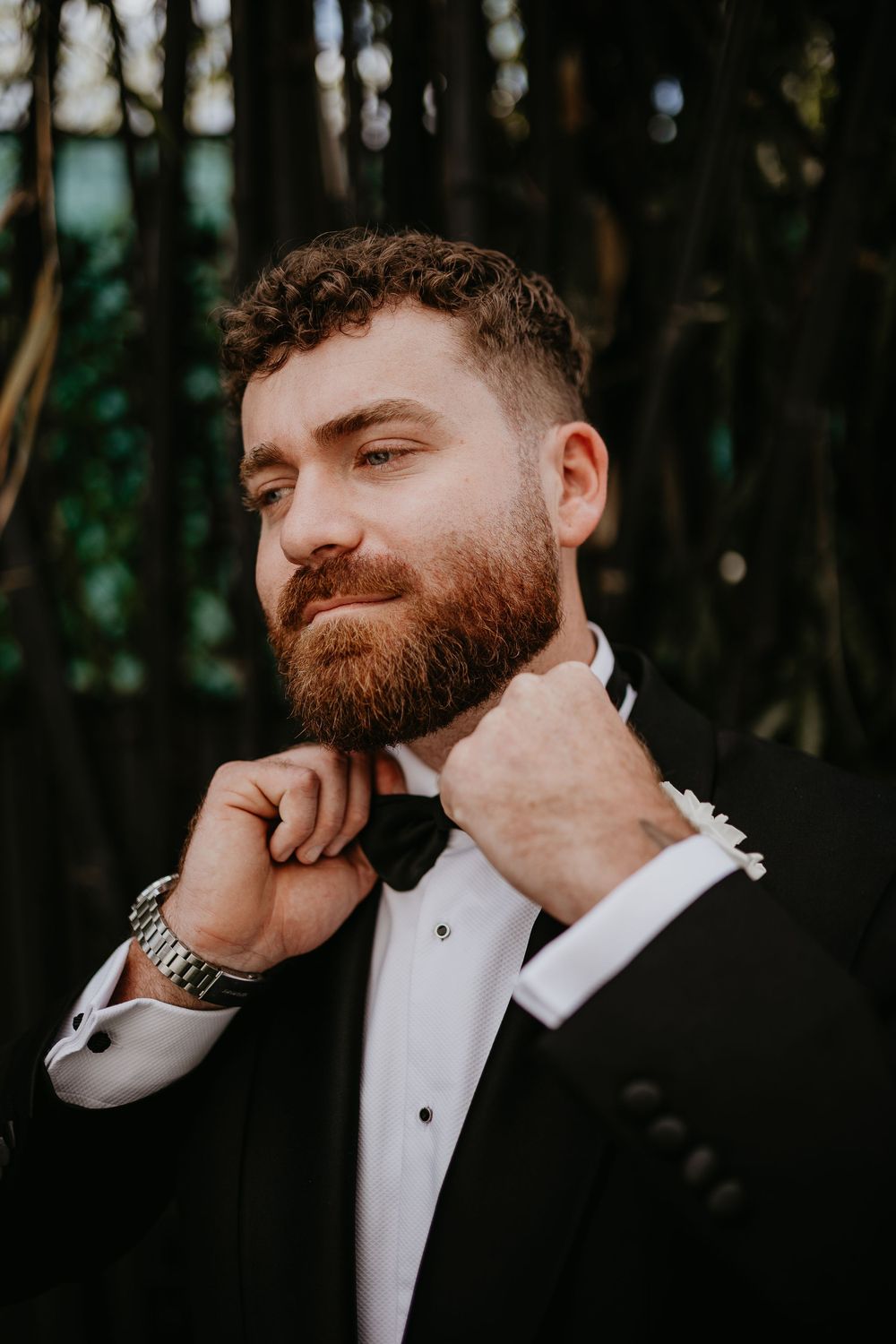 Groom in formal tuxedo and bowtie poses thoughtfully in moody forest setting.