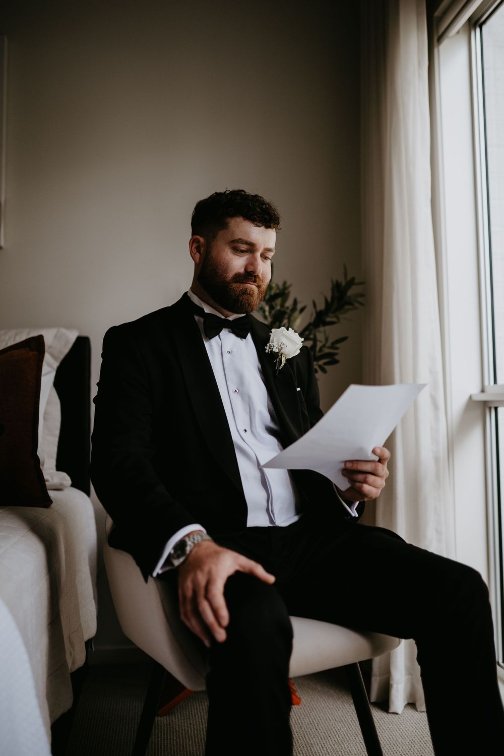 Groom in classic tuxedo reading letter while seated by window in natural light.