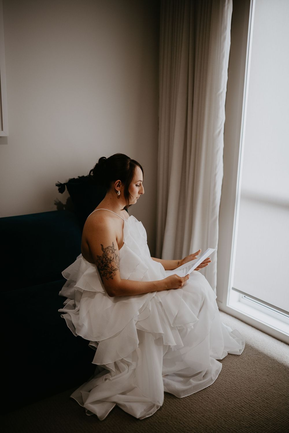Person in white wedding gown sits by window reading document in quiet moment before ceremony.