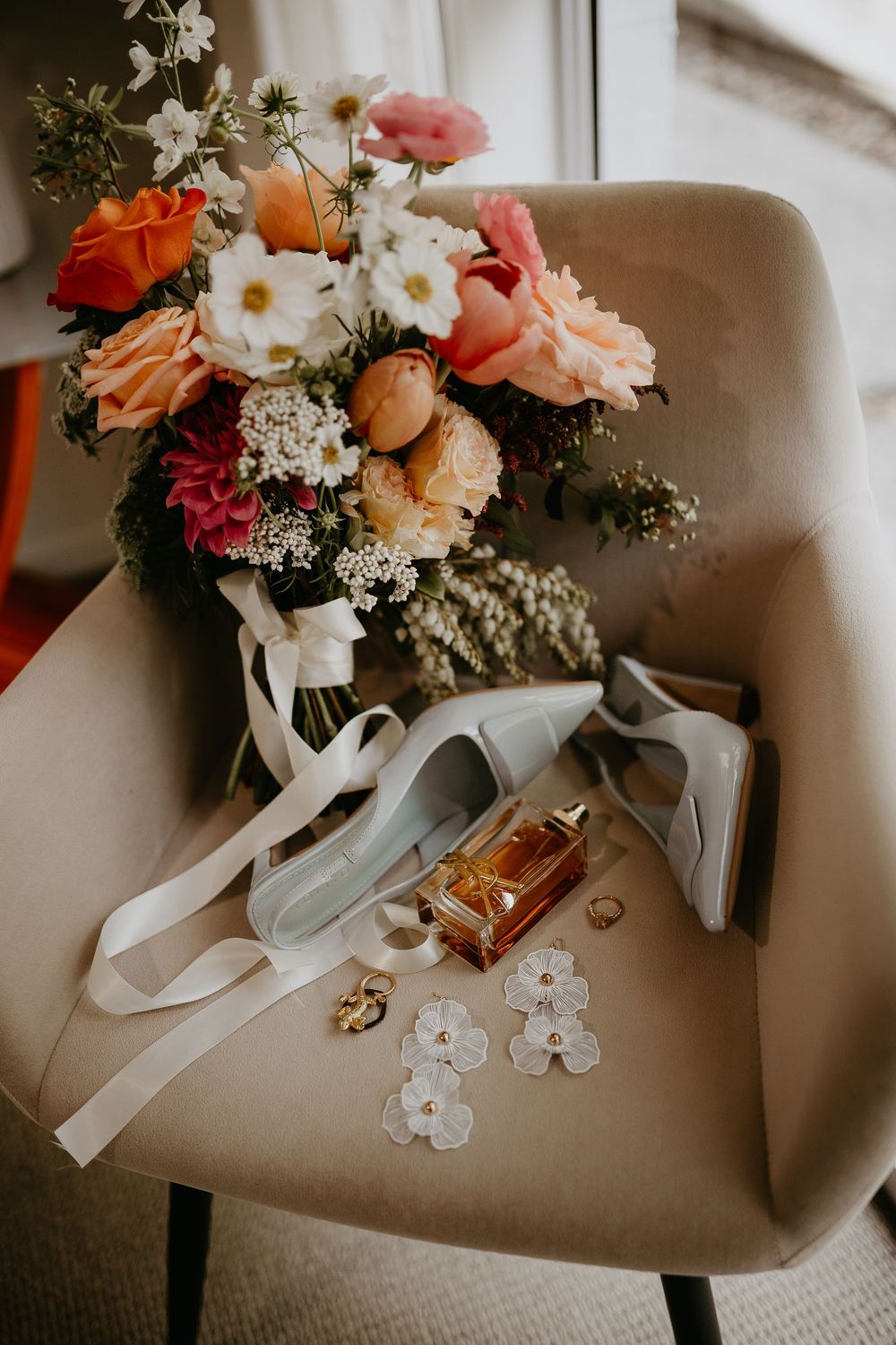 Wedding accessories including white floral bouquet, shoes, and jewelry arranged on a beige chair.